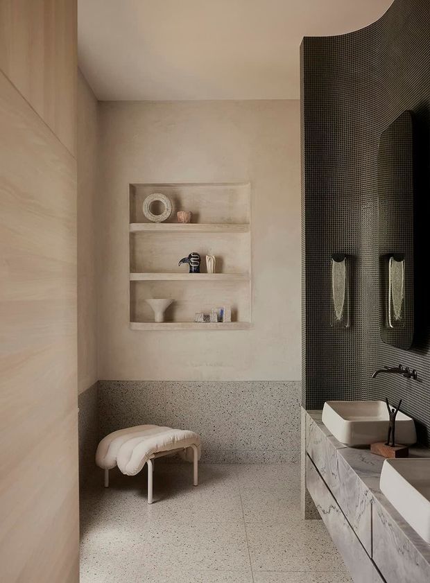 A refined contemporary bathroom in warm neutral tones, featuring a terrazzo floor and lower wall dado, pale timber wall panelling, integrated plaster shelving, and a dark small-format tiled curved feature wall. A long marble-look vanity with vessel basins, wall-mounted taps, mirrors and sculptural lighting occupies the right side, while a padded stool sits in the open area.