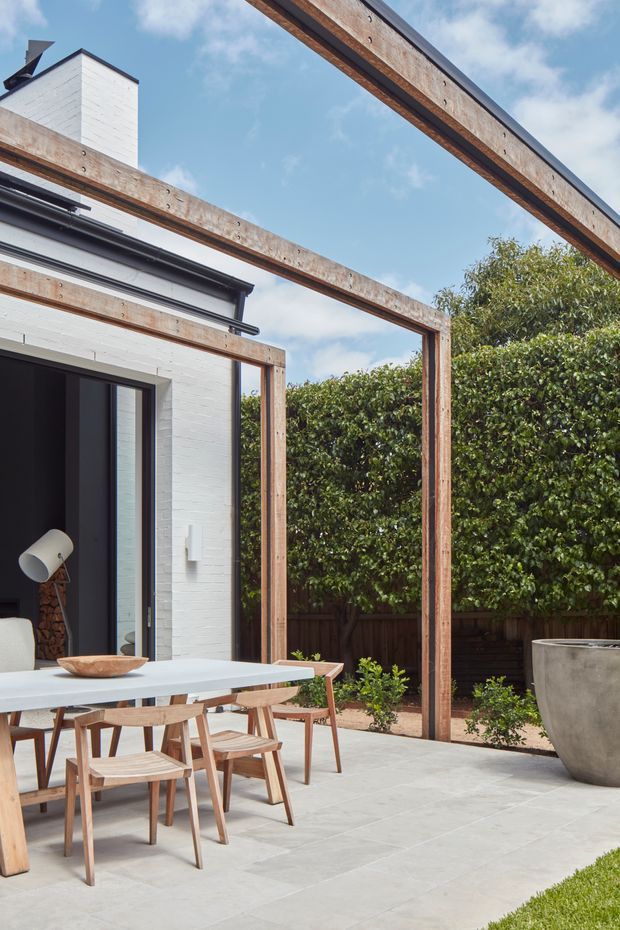 A contemporary outdoor dining terrace beside a white brick house, framed by tall timber pergola beams and posts. A pale dining table with timber chairs sits on large light-grey paved tiles, with a large concrete planter and dense hedging enclosing the courtyard. Black-framed glazing and a partially visible interior appear at the left.