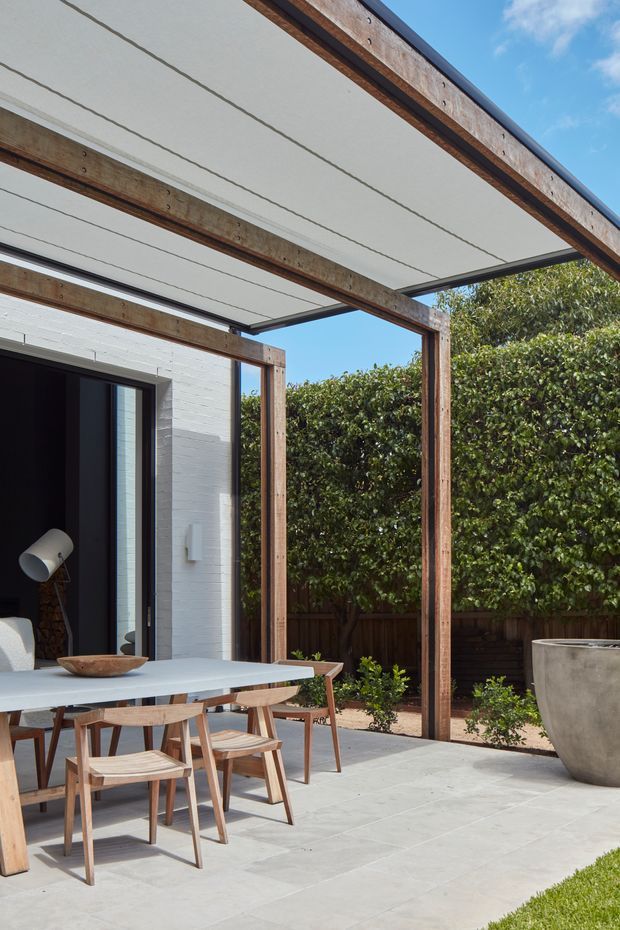 A bright contemporary outdoor dining terrace beneath a timber-framed pergola with a flat translucent or fabric-like roof canopy. The space features a large light-coloured dining table with timber chairs, pale large-format paving, a white brick exterior wall, dark-framed glazed doors, planted screening hedges, and a large concrete planter. The image is photographed in clear daylight and emphasizes the covered patio connection to the house.