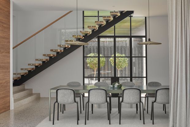 A refined open-plan dining area with a long glass-topped dining table and upholstered dining chairs, positioned beside a striking black steel floating staircase. Large black-framed industrial-style glazing fills the rear wall, while terrazzo stairs and flooring, pale walls, timber stair handrails, and pendant lights create a bright contemporary interior.
