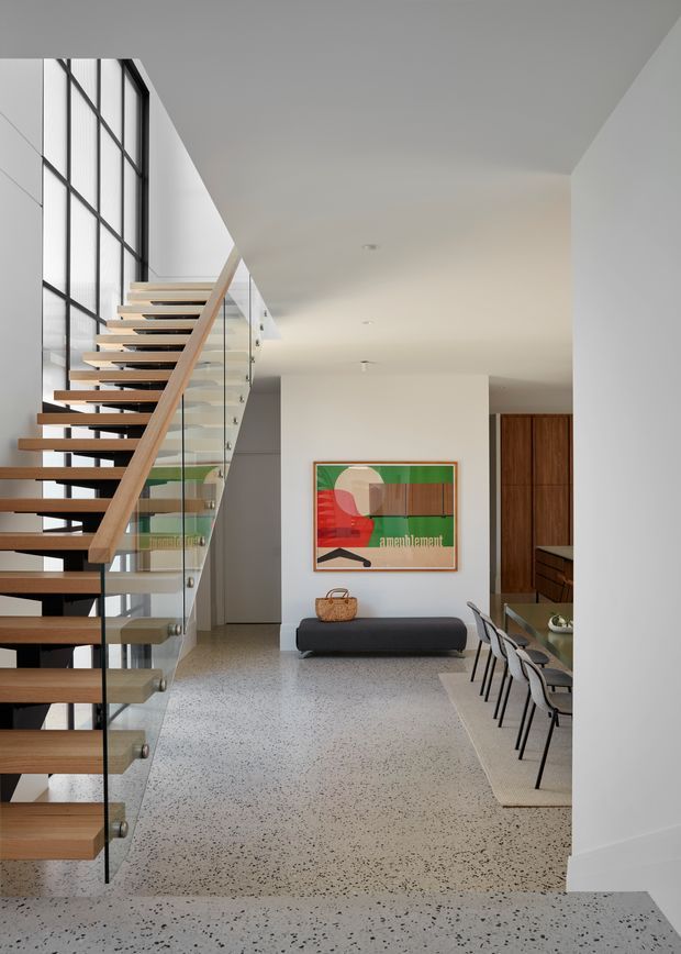 Bright contemporary open-plan entry and dining area featuring a floating timber staircase with glass balustrade, black metal-framed floor-to-ceiling glazing, polished terrazzo flooring, white walls and ceiling, and dark timber cabinetry. A compact bench and colourful framed artwork sit beneath the stair landing, while a dining table extends along the right side.