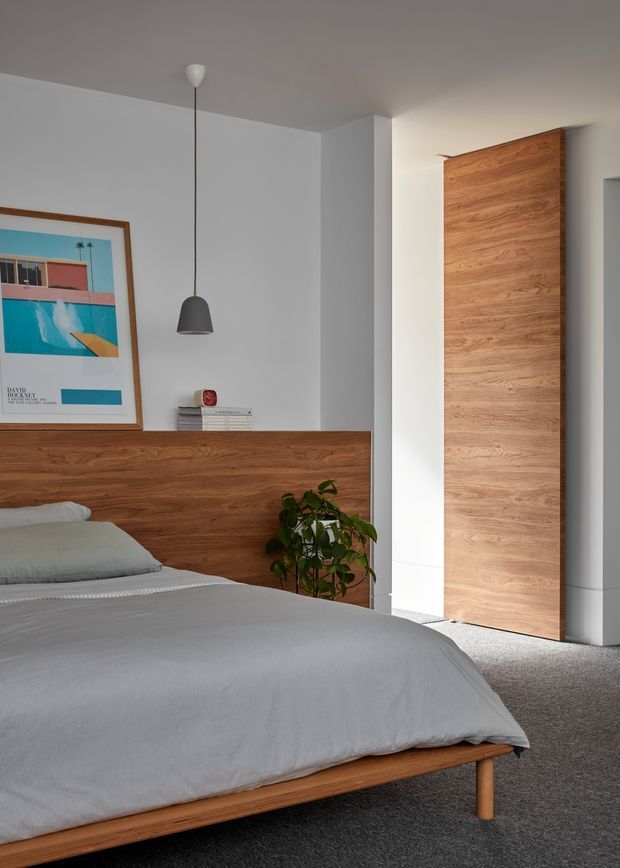A calm, contemporary bedroom with a low timber bed, grey bedding and carpet, a full-width timber headboard with integrated ledge, and a tall timber-clad wardrobe or door panel. White walls and ceiling provide a clean backdrop, complemented by a simple pendant light, framed artwork, and a small indoor plant.