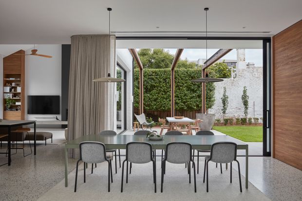 Contemporary open-plan dining area with a long dining table and upholstered chairs positioned beside full-height glazed doors. The space opens to a landscaped courtyard with a lawn, greenery, outdoor seating, and a white masonry boundary wall. Interior features include a polished light terrazzo or concrete-like floor, neutral curtains, dark-framed glazing, timber wall panelling, sculptural pendant lights, and a partially visible kitchen and living area.