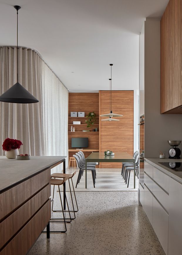 A refined open-plan kitchen and dining area with a warm timber island and cabinetry, pale stone or composite benchtops, and a terrazzo-style polished floor. The dining zone features a glass table, sculptural upholstered chairs, and a built-in timber shelving wall with a television and decor. Full-height sheer curtains soften the curved glazed wall, while minimalist black pendant lights provide contrast.