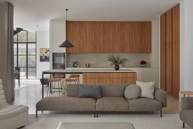 Contemporary open-plan living and kitchen area with a warm timber feature wall and cabinetry, pale stone or concrete-look splashback, central island with bar seating, and a large neutral grey sofa in the foreground. Floor-to-ceiling black-framed glazing opens toward a balcony or exterior, while a sculptural black pendant, recessed lighting, and minimal furnishings create a refined modern atmosphere.