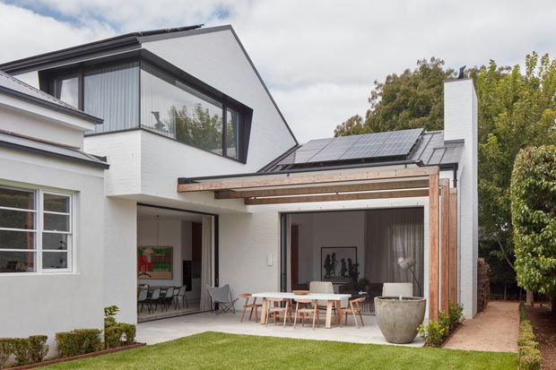 Exterior view of a contemporary residential extension with white rendered masonry walls, dark metal-framed glazing, a pitched roof, solar panels, and a timber pergola over an outdoor dining terrace. The design connects indoor dining and living spaces to a landscaped backyard, with a tall white chimney as a prominent feature.