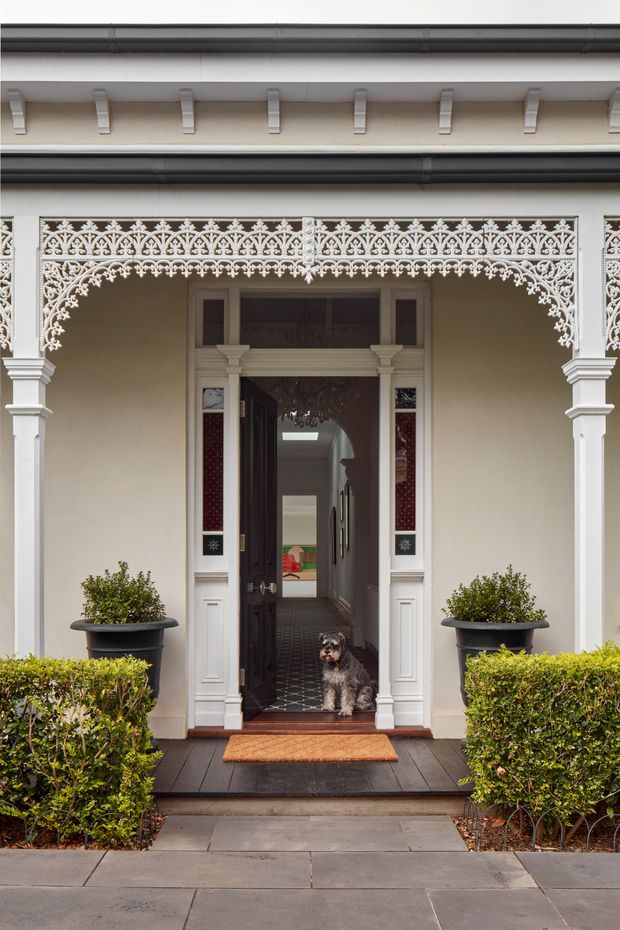 Front exterior view of an ornate heritage-style townhouse entry. The cream-painted facade features elaborate white lacework verandah detailing, decorative columns and brackets, dark guttering, a dark timber entrance door with sidelights, and a tiled hallway visible inside. Symmetrical clipped hedges and potted shrubs frame the entry, with a small dog standing in the doorway.