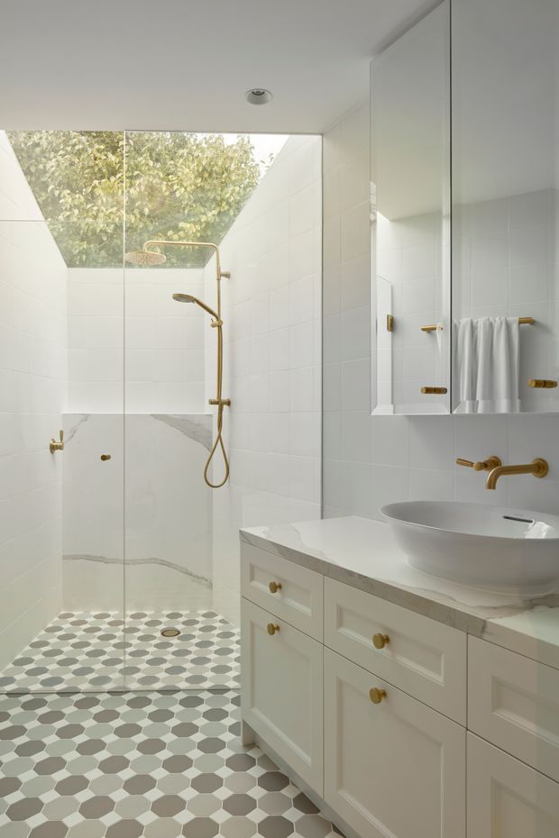 Bright, refined bathroom featuring a walk-in glass shower, white tiled walls, patterned hexagonal floor tiles, a marble-look vanity benchtop, white raised-panel cabinetry, and a vessel basin. Brushed brass/gold shower and tapware, towel rails, and cabinet hardware add warm accents. A high glazed opening brings in natural light and views of foliage.