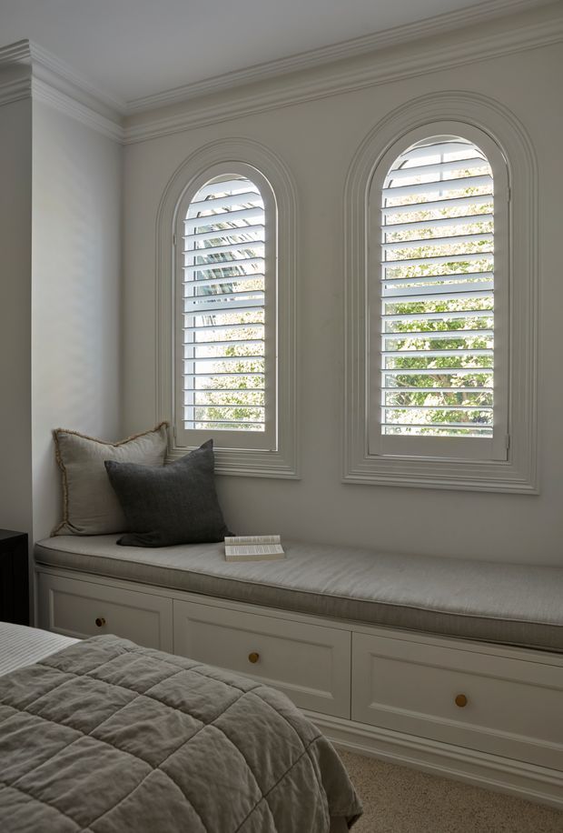 A serene, traditionally detailed bedroom corner featuring two tall arched windows with white plantation shutters, ornate layered mouldings, and a built-in upholstered window seat with drawer storage. A neatly made bed with a soft grey quilt occupies the foreground, while neutral textiles and a dark accent cushion create a calm, refined atmosphere.