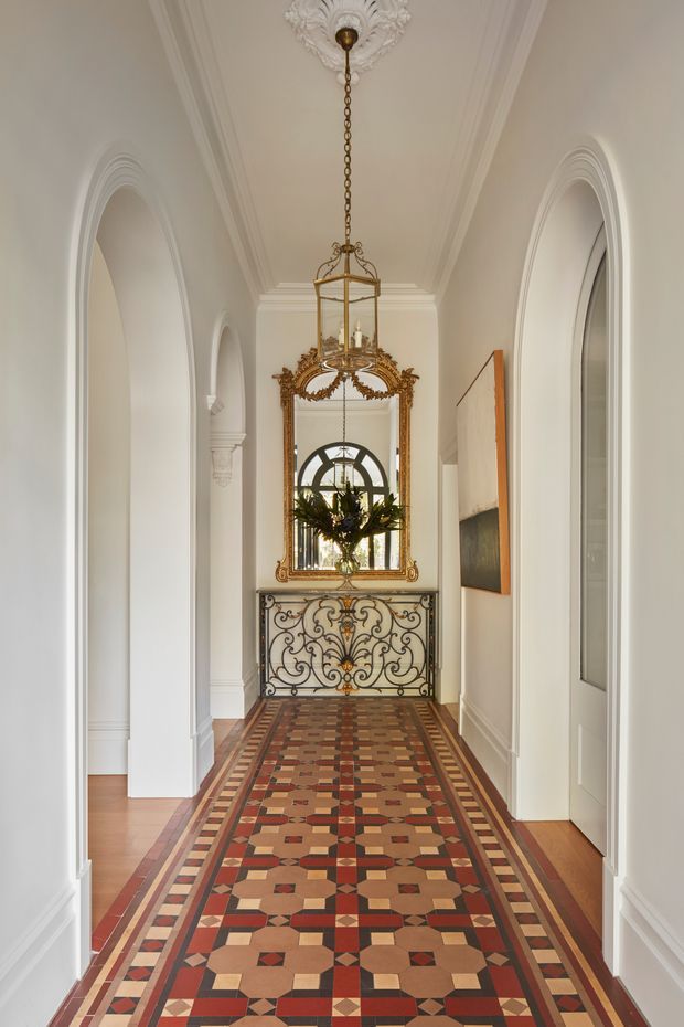 A refined residential hallway with white walls, tall arched openings, ornate decorative mouldings, and a patterned geometric tiled floor runner. A large elaborately carved gilt mirror and decorative console table form a focal point at the end of the corridor, complemented by a suspended brass lantern and a floral arrangement. The space has strong heritage detailing with a formal, elegant character.