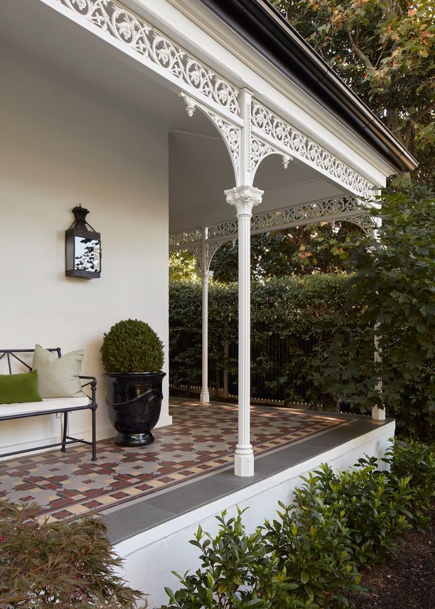 A covered front veranda or porch with ornate white cast-iron decorative columns, lacework brackets and friezes. The veranda has a patterned geometric tiled floor, white walls, a black decorative wall lantern, a simple metal bench with cushions, and a large black planter. Dense hedging and garden planting surround the elevated porch, creating a refined heritage-style entry exterior.