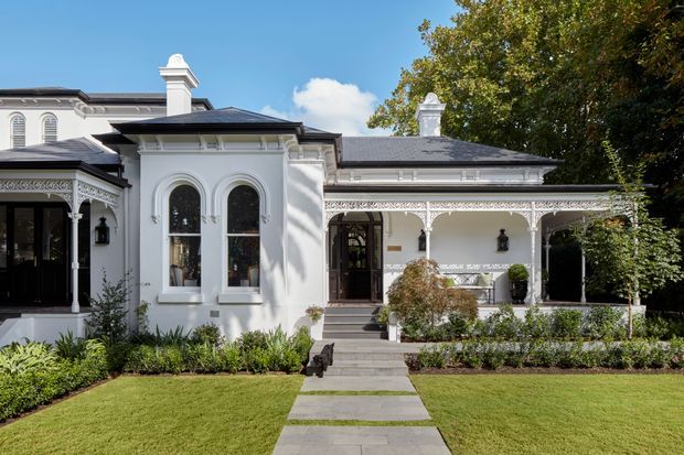 A grand white heritage-style residence viewed from the front garden, featuring ornate architectural detailing, arched windows, decorative veranda fretwork, classical chimneys, dark hipped roofing, and a formal paved pathway through manicured landscaping.