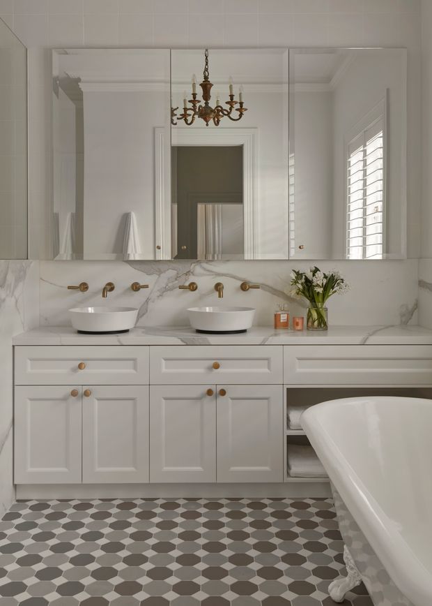 A refined traditional bathroom featuring a double vanity with two white vessel basins, marble-look wall and benchtop surfaces, mirrored cabinet doors, brass wall-mounted tapware, panelled white cabinetry, and a freestanding white bathtub. A geometric monochrome patterned floor, plantation shutters, and an ornate brass chandelier add classic character.