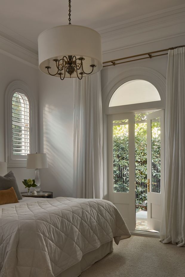 Elegant bedroom with a calm, light-toned palette. The room features a neatly made bed with a quilted white coverlet, layered neutral cushions, a small bedside table and lamp, tall white curtains, ornate arched windows and glazed double doors opening to a leafy garden. Decorative ceiling cornices, curved window detailing, and a central drum pendant with traditional metalwork add refined character.