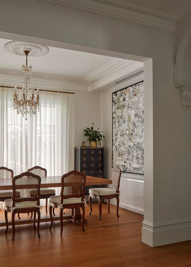 Elegant formal dining room viewed through a wide cased opening, featuring a long polished timber dining table, matching cane-back timber chairs, a dark traditional chest of drawers, sheer full-height curtains, ornate crystal chandelier, decorative ceiling medallion and detailed plaster cornices. A large textured artwork sits above a radiator, while the warm timber floor and restrained neutral walls create a refined, classic atmosphere.