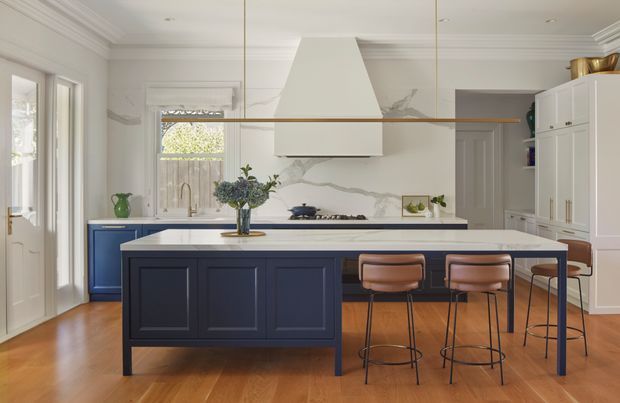 A bright, refined kitchen with a large navy-blue island, white stone benchtop, timber flooring, white cabinetry, marble-look splashback, and a sculptural white rangehood. Four leather bar stools line the island, while a long timber shelf and brass-toned details add warmth. The room features classic wall panelling, decorative ceiling cornices, and generous glazing.