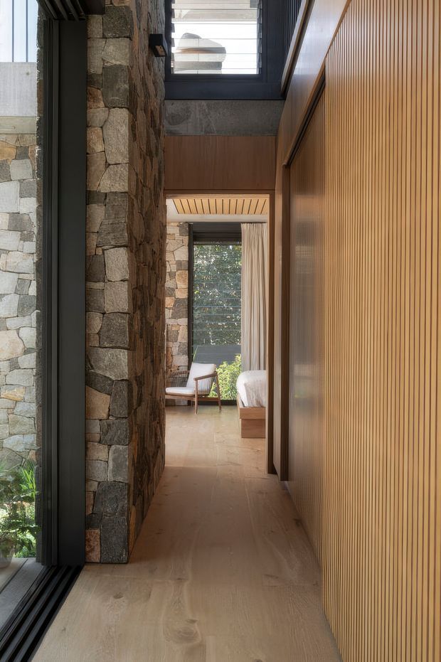 A refined interior passage connects to a bright sitting area. The corridor features a warm timber floor, a full-height vertical timber slat wall, exposed rugged stonework, dark metal-framed glazing, and a timber-lined ceiling detail. Through the opening, upholstered lounge chairs and a sofa are visible beside large windows with curtains and garden views. The design combines natural stone and timber with strong vertical lines and abundant daylight.