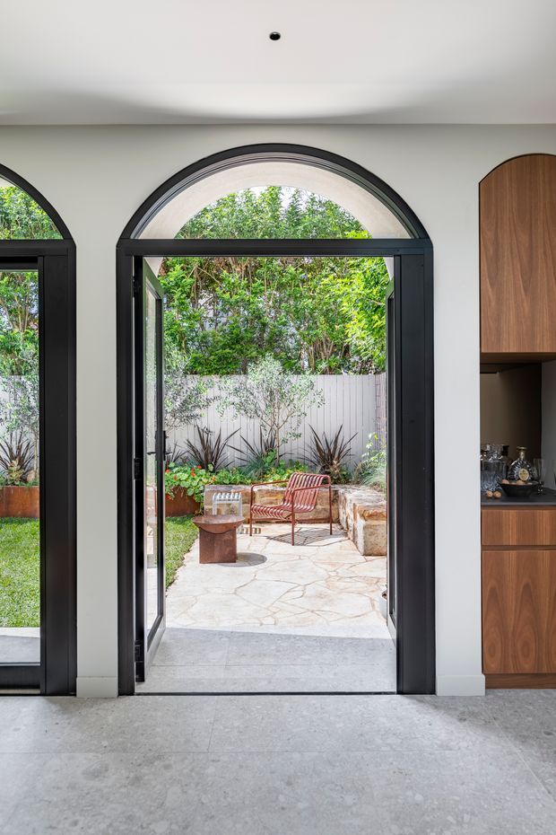 A bright contemporary interior looking through a tall arched black-framed glazed doorway into a landscaped courtyard. The room has a pale grey polished concrete floor, smooth light walls, and a built-in dark timber cabinet with an arched upper profile on the right. Outside is a stone-paved seating area with a bench, chairs, planters, and dense greenery. Matching arched glazing is partially visible on the left.