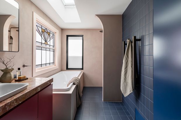 A stylish contemporary bathroom viewed from the vanity area, featuring a freestanding bathtub beneath a decorative leaded-glass window, a skylight, warm plaster walls, dark blue tiled walls and floor, and a dark reddish wood vanity with a stone benchtop. Black-framed glazing, black tapware and towel rails provide contrast.