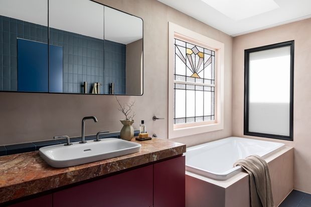 A refined contemporary bathroom featuring a wide white vessel basin on a dark red built-in vanity with a richly patterned stone benchtop. The room has warm taupe walls, dark blue tiled surfaces reflected in the mirror, a freestanding-style white bathtub, black-framed frosted glazing, and a decorative leadlight-style window with geometric coloured glass. The palette is warm and sophisticated with burgundy, taupe, dark blue, black, and white accents.