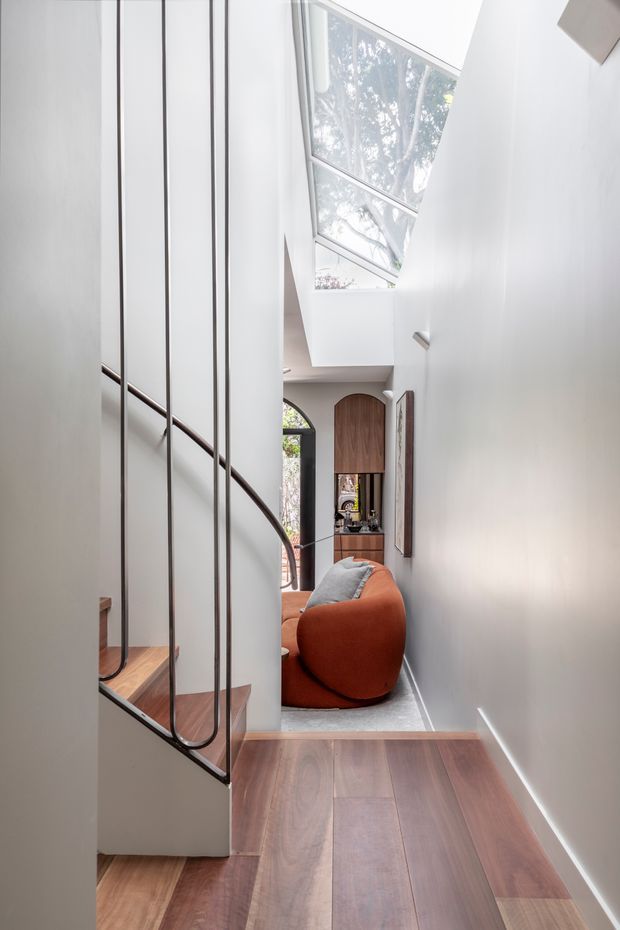 A bright, narrow interior hallway beside a sculptural staircase. The stair has warm timber treads and a slender dark metal balustrade, while a large overhead skylight and clerestory glazing bring in abundant natural light. A compact living area with a curved rust-orange lounge chair and grey cushion is visible at the end, along with curved timber cabinetry and a small arched window.