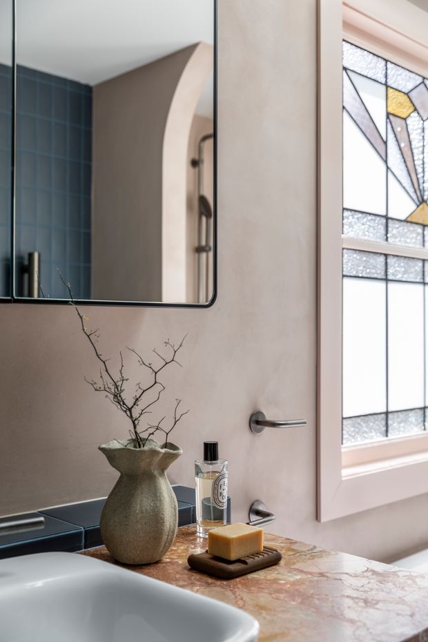 Close-up view of a refined bathroom vanity with a pale stone vessel sink, warm marble countertop, matte plastered walls, dark blue wall tiles, and a frameless rounded-corner mirror. A sculptural ceramic vase, toiletries, and soap sit on the counter. A decorative geometric stained-glass window with pale pink trim adds an Art Deco-inspired feature, while brushed metal tapware and a curved doorway are visible in the mirror reflection.