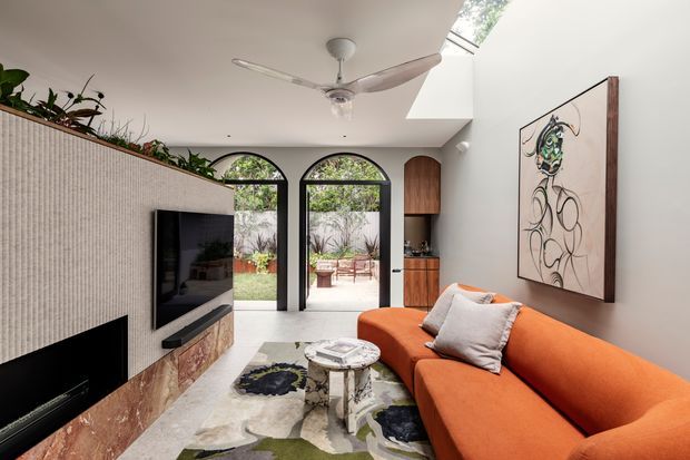A bright contemporary living room with a large orange sectional sofa, wall-mounted television, linear fireplace, and a textured light wall finish. Arched glazed doors open to an outdoor patio, while a skylight and ceiling fan add natural light and ventilation. Built-in timber cabinetry and a stone-clad fireplace surround complete the space, with large abstract artwork and indoor plants providing decorative accents.
