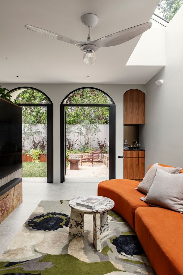 A bright contemporary living room with a large orange sectional sofa, patterned area rug, sculptural marble coffee table, and a wall-mounted television. Black-framed arched glazed doors open to a landscaped courtyard, while a compact timber bar or kitchenette is built into the side wall. A ceiling fan and skylight enhance the light-filled space.