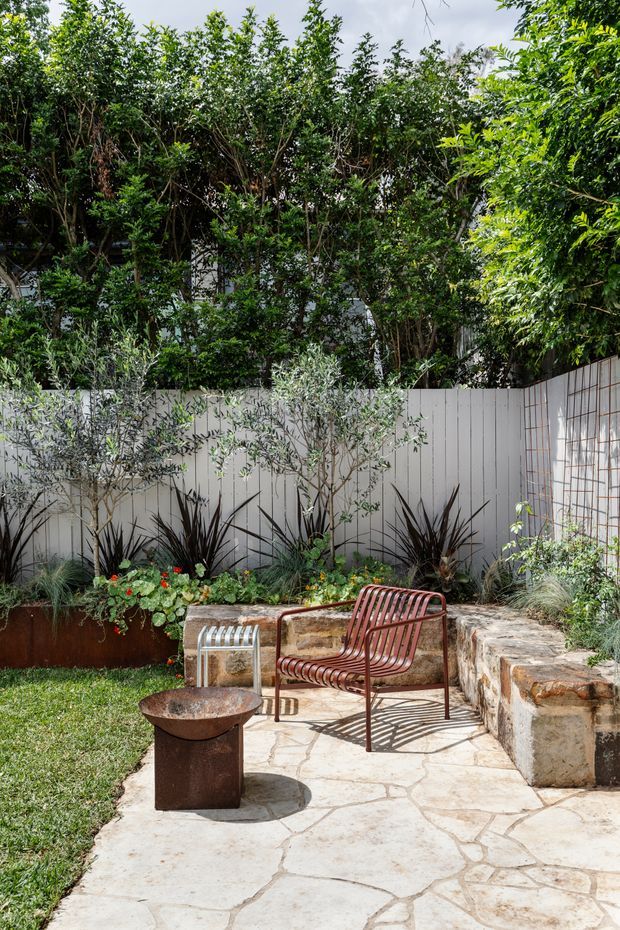 A landscaped outdoor courtyard or garden seating area with an irregular pale stone-paved patio, low rustic stone retaining wall, white vertical-panel privacy fencing, and dense planting. A rust-coloured slatted metal lounge chair sits beside a small weathered metal fire bowl, with a raised planted bed and additional garden furniture nearby. The scene is bright, natural, and relaxed.