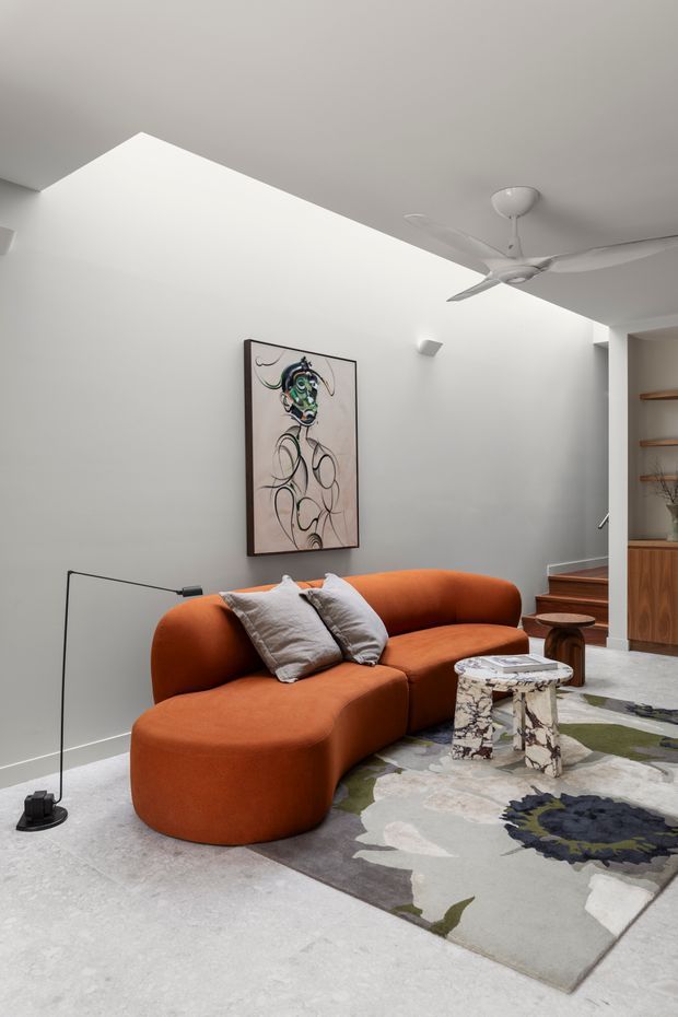 A bright contemporary living room with white walls and a high angled ceiling, centred around a large curved rust-orange sectional sofa. A sculptural patterned rug, marble-look coffee table, slim black floor lamp, abstract wall artwork, timber stair treads, and built-in timber shelving complete the space. A white ceiling fan and recessed lighting provide functional illumination and ventilation.