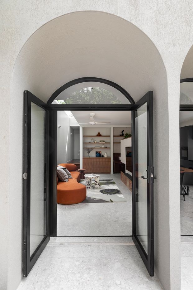 View through open black-framed glazed double doors beneath a large arched opening into a refined living room. The room features a curved rust-orange sofa, patterned area rug, low stone coffee table, timber built-in cabinetry with open shelving, and a white ceiling fan. Light textured plaster walls and pale concrete-like flooring create a bright, contemporary interior, while a mirrored or glazed opening at right provides a partial reflection of adjacent joinery.