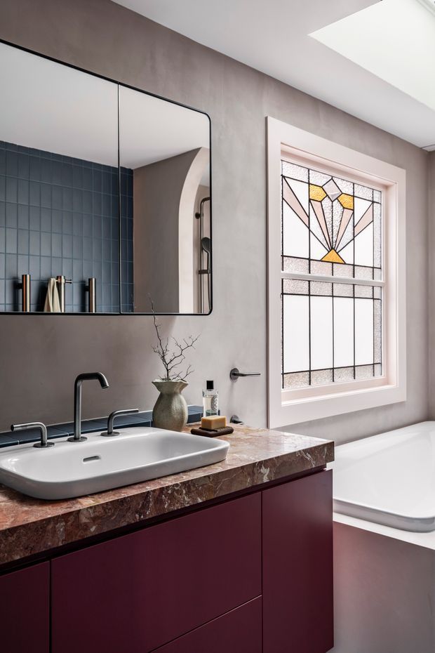 A refined bathroom featuring a large frameless mirror, a white vessel-style basin with dark metal tapware, a burgundy built-in vanity, and a richly veined stone benchtop. The walls are finished in a warm grey plaster, with dark blue vertical wall tiles behind the basin. A white bathtub sits beneath a decorative geometric stained-glass window, and a skylight adds natural light. The overall look is contemporary with art deco-inspired detailing.