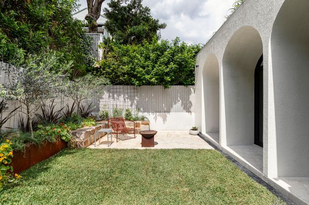 A bright landscaped courtyard beside a white rendered building with a series of tall arched openings and dark-framed doors. The outdoor area includes a small lawn, paved patio, raised rusted-metal garden bed, layered planting, outdoor chairs, a low fire bowl, and a pale slatted privacy fence. The scene has a refined contemporary Mediterranean feel with strong indoor-outdoor connection.