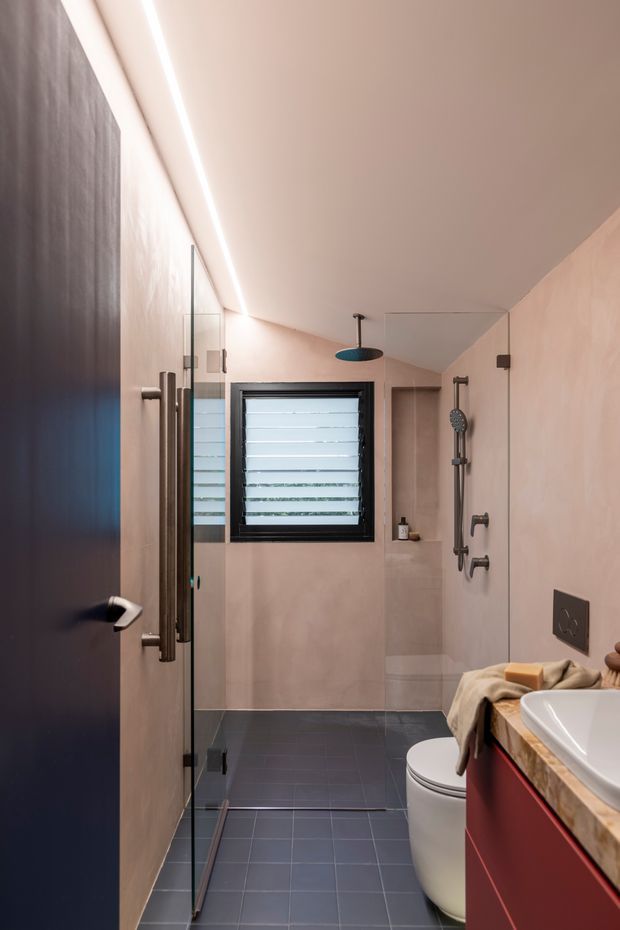 A narrow contemporary bathroom with a walk-in glass shower, wall-mounted rain shower and hand shower, toilet, and red vanity with a stone-look benchtop. The room has dark tiled flooring, warm blush-toned wall finishes, black-framed louvred window, integrated linear ceiling lighting, and dark hardware. A partially visible dark door opens into the bathroom.
