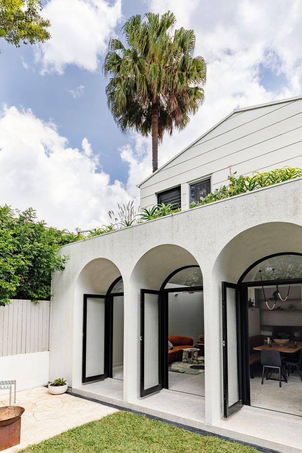 Bright exterior view of a white rendered courtyard extension with a row of repeated rounded arches and black-framed glazed doors, some open to reveal a contemporary living and dining interior. A planted terrace sits above the arches, with a tall palm tree and garden paving in the surrounding courtyard.