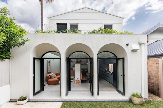 Exterior view of a compact two-storey residential extension or rear facade. The white rendered lower level features three tall round-arched openings with black-framed glazed folding or hinged doors, opening onto a small paved area and lawn. Interior spaces are visible through the openings, including a furnished living area and a dark kitchen or joinery zone. The upper level has a simple white weatherboard-style gabled facade with horizontal window openings. Potted plants and neighbouring residential structures frame the scene.