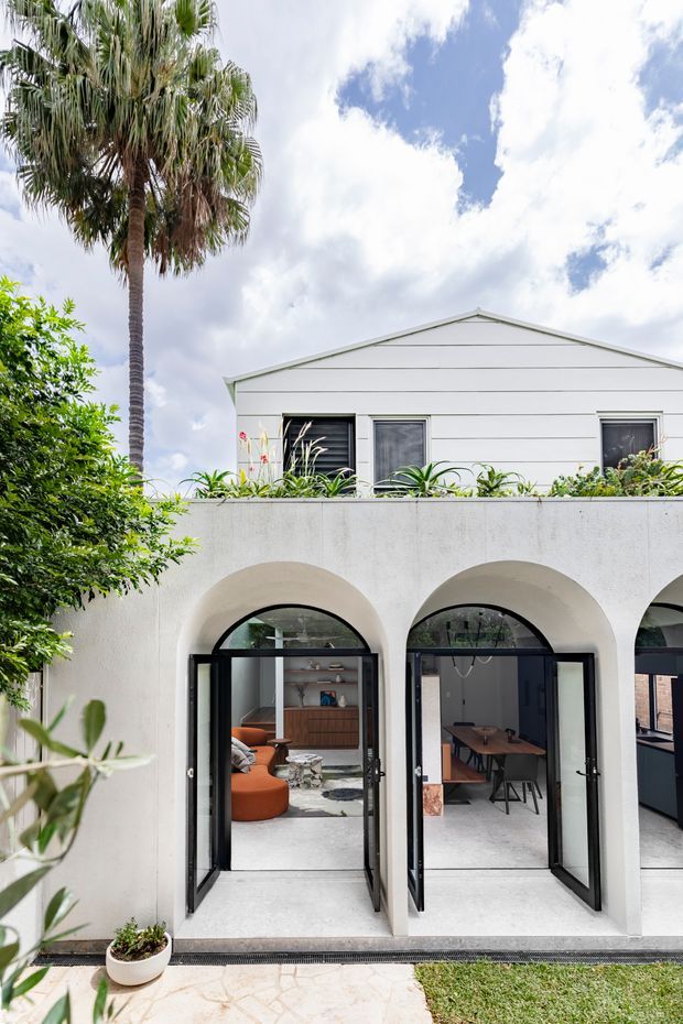 Bright exterior view of a two-storey residence with a white rendered lower facade featuring repeated rounded arch openings and black-framed glazed folding doors. Through the open doors, a contemporary living and dining area is visible. The upper level has white horizontal weatherboard cladding and a simple gable roof, with tropical planting and a tall palm tree framing the property.