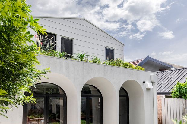 Exterior view of a light-coloured, two-storey residential building with a simple pitched gable roof, horizontal cladding on the upper level, and a distinctive ground-floor arcade of rounded arches framing dark glazed doors and windows. Planter greenery lines the upper terrace and trees partially frame the facade. Adjacent roof and brickwork are visible in the background.