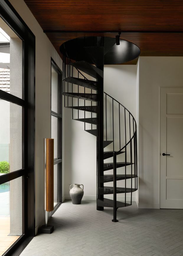 A refined interior hallway or entry space featuring a sculptural black metal spiral staircase with perforated treads and a curved railing. Full-height black-framed glazing lines the left side, while a warm timber slatted ceiling contrasts with pale walls and a light grey herringbone-patterned floor. A simple panelled door, sculptural ceramic vessel, and slim wall-mounted timber feature add understated detail.