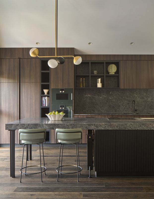 A sophisticated contemporary kitchen viewed from the living side of a large island. The room features dark timber cabinetry, a dark stone island benchtop and splashback, integrated appliances, open display shelving, a ribbed dark island base, and two upholstered metal-framed bar stools. A sculptural multi-globe pendant adds a small brass, white, black, and gold accent above the island. Wide timber flooring and a low white ceiling complete the refined, moody interior.