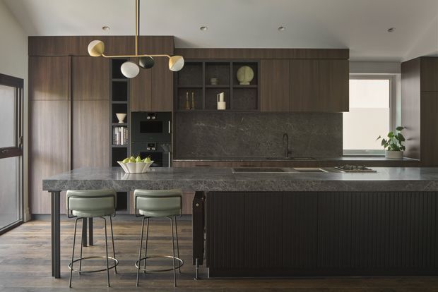 A sophisticated contemporary kitchen with dark timber cabinetry, a large charcoal stone island and matching stone splashback. The island includes seating for two with upholstered stools, while integrated appliances and open shelving are set into the full-height joinery. A sculptural multi-globe pendant light hangs above the island, and natural light enters through the glazed door and window.
