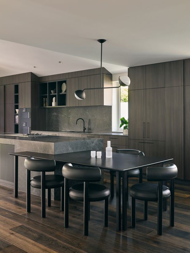 A sophisticated open-plan kitchen and dining area with extensive dark timber cabinetry, a large stone island, integrated open shelving, and a dark dining table surrounded by black upholstered chairs. The room has dark timber flooring, a pale ceiling, a narrow window bringing in daylight, and a sculptural linear pendant light with globe shades. The overall look is refined, moody, and contemporary.