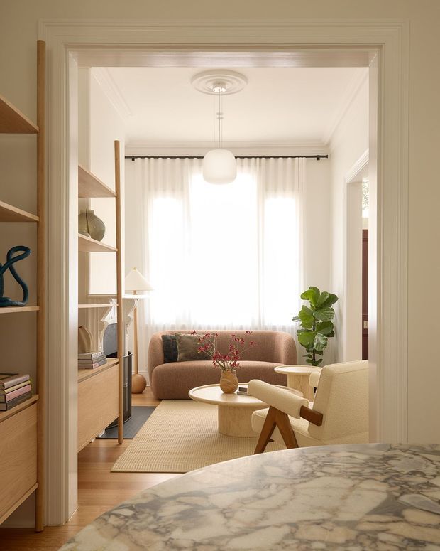 A warm, refined living room viewed through a wide trimmed doorway from an adjacent space. The room features a caramel upholstered sofa, cream lounge chair, round light-wood coffee table, textured neutral rug, sheer floor-to-ceiling curtains, built-in open shelving and cabinetry, timber flooring, a central globe pendant, and a leafy indoor plant. A marble-patterned benchtop or table surface occupies the foreground, with classic wall and ceiling mouldings framing the opening.