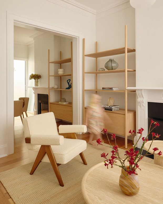 Warm, refined living room with cream walls, ornate white ceiling cornices and fireplace detailing, light timber built-in open shelving, and a textured neutral rug. A sculptural cream boucle lounge chair sits beside a rounded light-wood coffee table with red floral branches. A dining area and fireplace are visible through the wide opening and mirror-like reflections.
