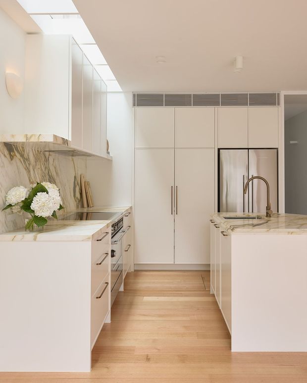 Bright, refined contemporary kitchen with a galley-style layout and central circulation aisle. The space features white flat-panel cabinetry, integrated tall storage, stainless-steel appliances, pale veined marble benchtops and splashback, a brass-toned tap, and light timber flooring. A skylit ceiling and concealed linear ventilation add to the clean, airy design.