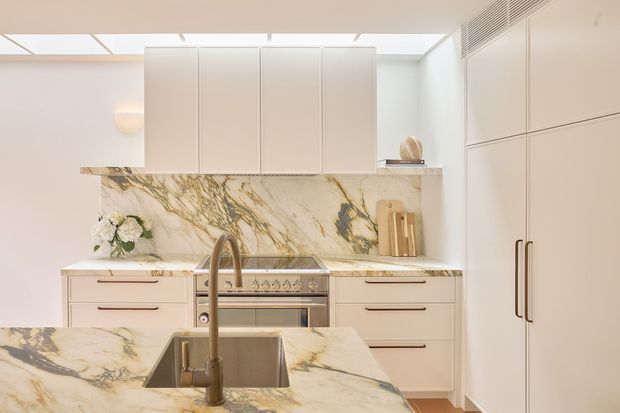 Bright, refined kitchen with pale cabinetry, a large marble island and matching marble splashback and benchtops. The space includes a double-bowl sink with brushed metal tapware, a freestanding-style range, integrated tall storage, warm timber flooring and a linear ceiling skylight or clerestory element.