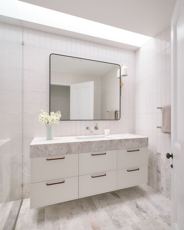 Bright, refined bathroom featuring pale tiled walls and a matching light stone-look floor, a floating double vanity with white drawers and slim bronze handles, an integrated basin, a large rounded-corner black-framed mirror, wall-mounted lighting, and a linear skylight or ceiling opening. A partially visible bathtub appears at left, while towels and a door are visible at right.