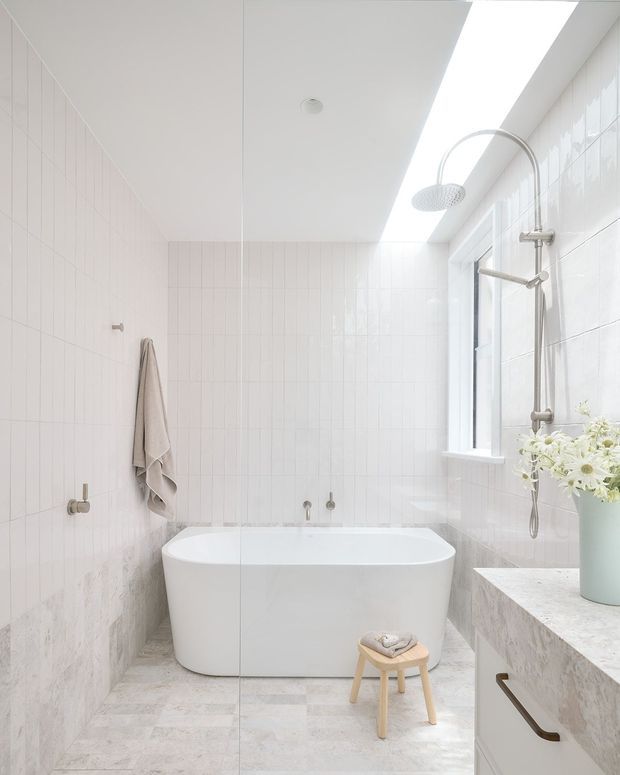 Bright, minimalist bathroom featuring a freestanding white bathtub beneath a large skylight, a wall-mounted rain shower, pale vertically stacked wall tiles, light marble-look floor and vanity surfaces, and a narrow window. A glass shower screen divides the bath area, while a small timber stool and pale vase add subtle warmth.