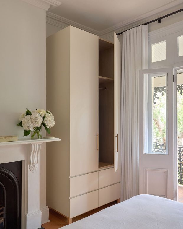 A refined bedroom interior featuring a tall built-in wardrobe with cream cabinetry, open timber-toned shelving, drawers, and brass-toned handles. A decorative white fireplace mantel with floral arrangement sits to the left, while a glazed French door with sheer curtains brings in natural light. Ornate ceiling cornices, wall mouldings, timber flooring, and a partially visible bed create a classic, elegant atmosphere.