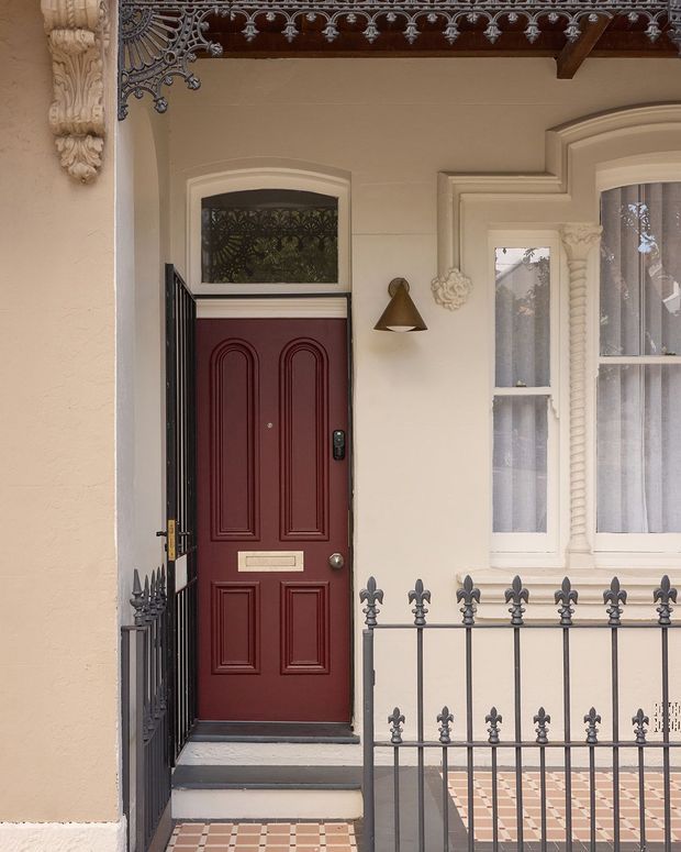 Close-up view of a decorative residential entry facade featuring a deep burgundy paneled front door with brass mail slot and hardware, a black wrought-iron gate and railing, cream rendered walls, ornate plaster detailing around the windows and eaves, and patterned tiled paving.